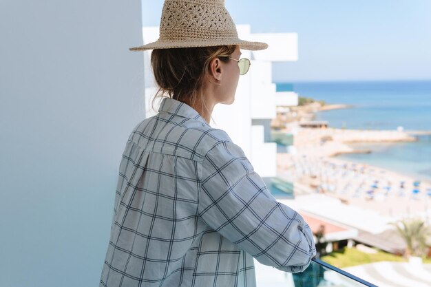 Une jeune femme portant un chapeau de paille profite de la vue depuis le balcon d'un hôtel ou d'un appartement en bord de mer