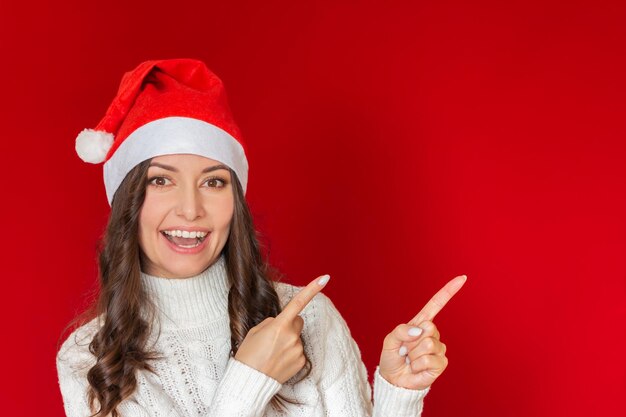 une jeune femme portant un chapeau de Père Noël sourit et pointe ses doigts sur un fond vide Copier l'espace