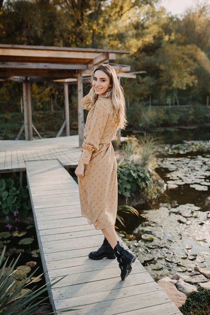 Jeune femme portant une longue robe en mousseline de soie marchant sur un pont en bois au bord d'un lac en souriant
