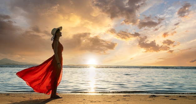 Jeune femme portant une longue robe rouge et un chapeau de paille debout sur la plage de sable au bord de la mer