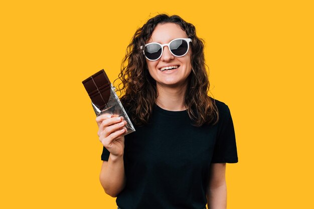 Photo une jeune femme portant des lunettes de soleil sourit et tient une barre de chocolat noir sur fond jaune