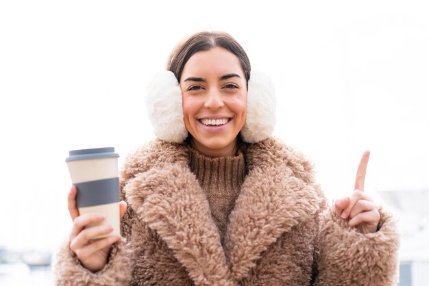 Photo jeune femme portant des manchons d'hiver et tenant du café à emporter à l'extérieur pointant vers le haut une excellente idée