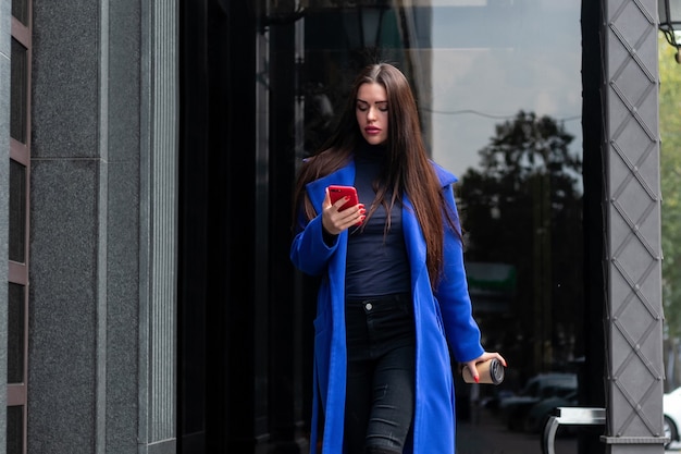 Une jeune femme portant un manteau bleu boit du café pour aller parler au téléphone dans la rue. Tir moyen verrouillé en temps réel