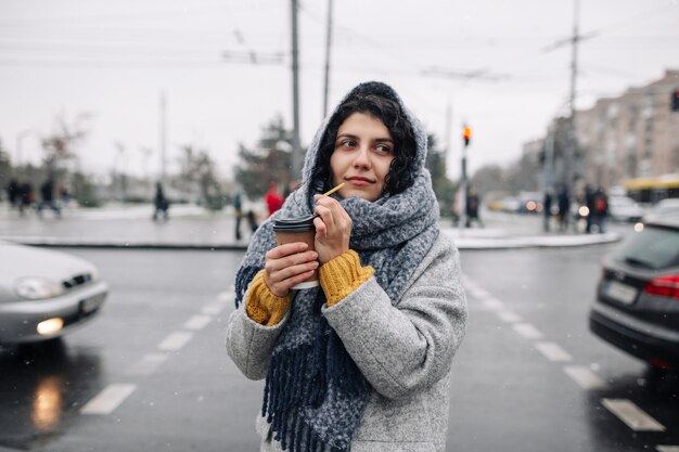 Jeune femme portant un manteau gris et une écharpe bleue se dresse sur un passage pour piétons avec une tasse de papier de café dans ses mains.