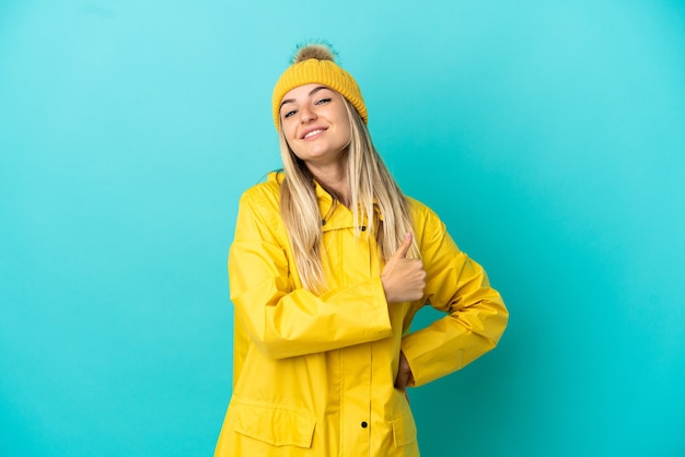 Jeune femme portant un manteau imperméable sur fond bleu isolé donnant un coup de pouce geste