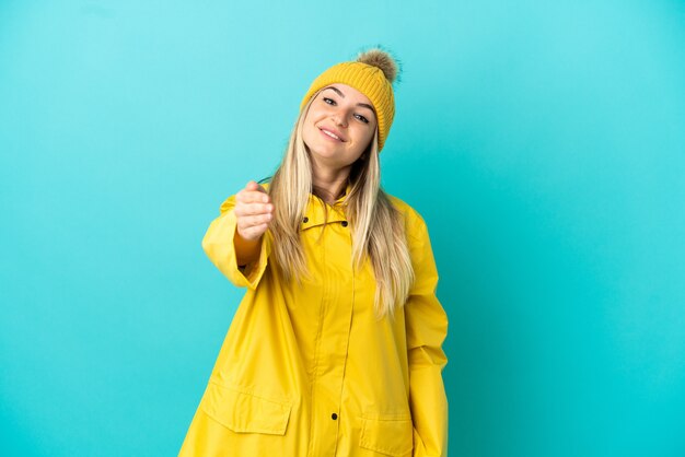 Jeune femme portant un manteau imperméable sur fond bleu isolé se serrant la main pour conclure une bonne affaire