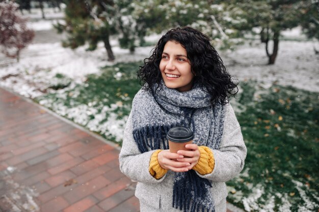 Jeune femme portant un manteau de mode frey et une écharpe bleue se dresse avec un café pour aller dans un parc enneigé d'hiver.