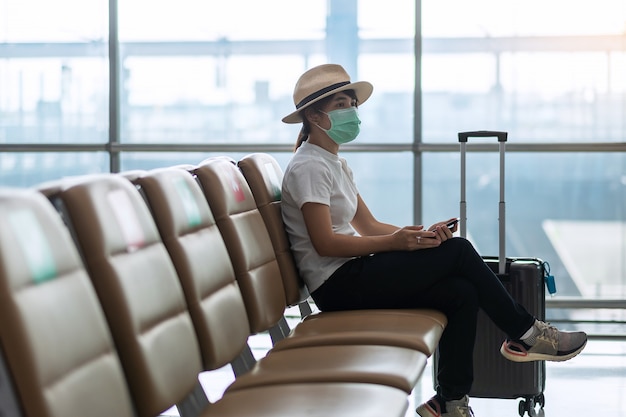 Jeune femme portant un masque facial et assise sur une chaise à l'aéroport, protection contre la maladie à coronavirus (Covid-19), femme asiatique voyageur. New Normal, bulle de voyage et distanciation sociale