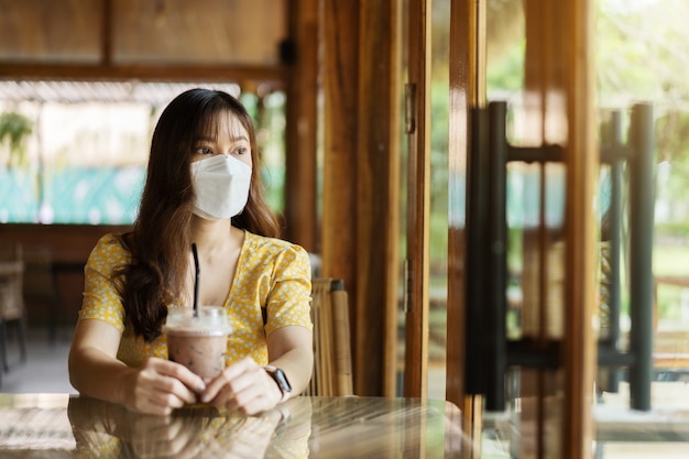 Jeune femme portant un masque facial pour la protection contre le coronavirus (covid-19) et buvant du lait au chocolat au café