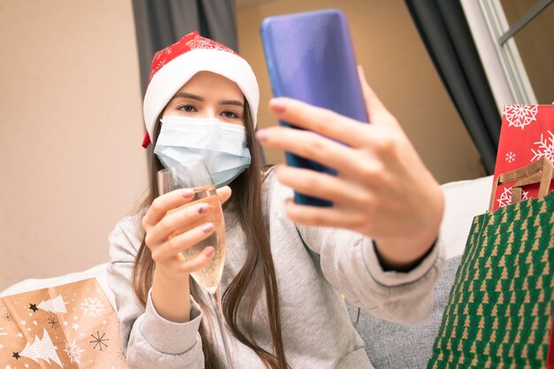 Une jeune femme portant un masque médical et un bonnet de Noel à la maison félicite ses amis pour Noël et le nouvel an via un smartphone, le père Noël avec une coupe de champagne