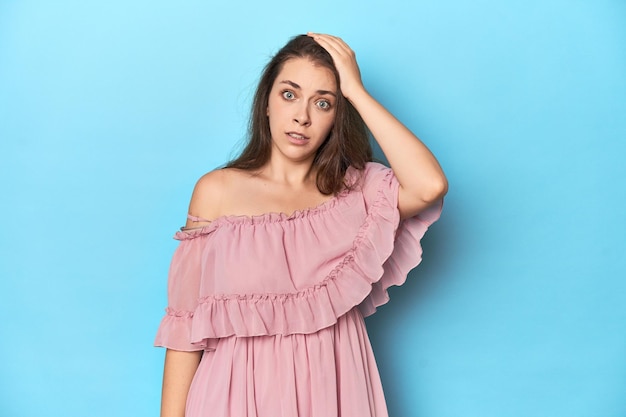 Une jeune femme portant une robe rose sur un fond de studio bleu est choquée