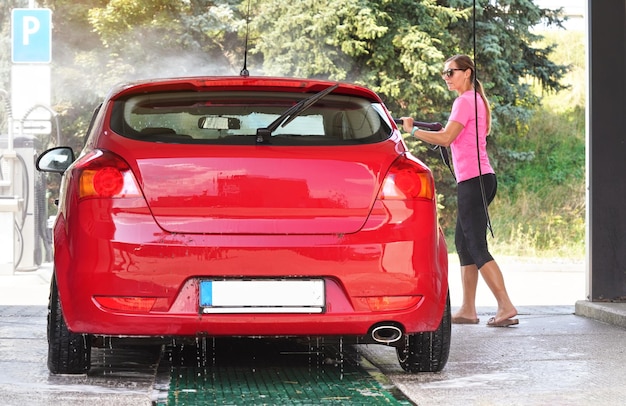 Jeune femme portant un t-shirt rose et des lunettes de soleil lavant sa voiture rouge dans un lave-auto en libre-service, vue de l'arrière