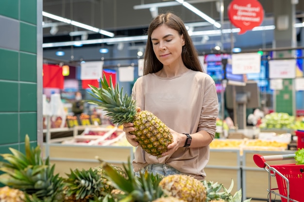 Jeune femme portant une tenue décontractée choisissant le meilleur ananas dans un supermarché moderne