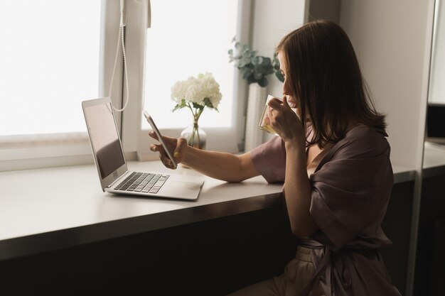 Une jeune femme porte des vêtements de couchage travaillant sur un ordinateur portable tout en étant assise dans le salon et en buvant du thé sur le réseau social et en travaillant à la maison