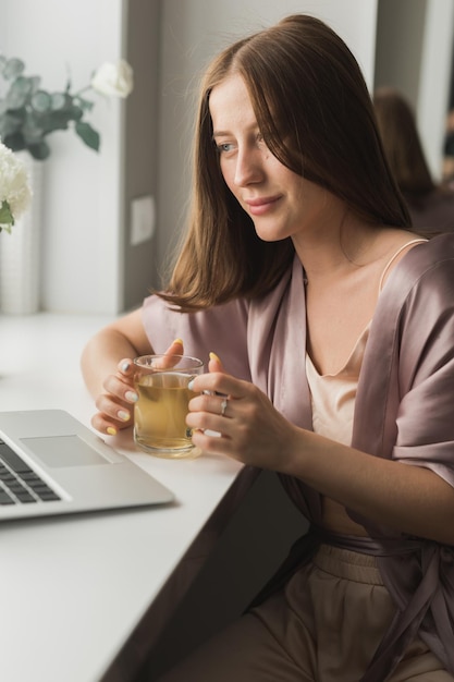 Une jeune femme porte des vêtements de couchage travaillant sur un ordinateur portable tout en étant assise dans le salon et en buvant du thé sur le réseau social et en travaillant à la maison