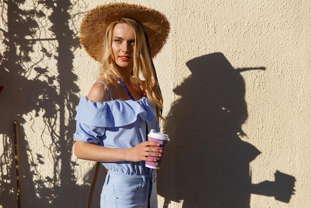 Jeune femme portée à la mode au chapeau de paille avec sac et tasse de café à emporter debout au café en plein air