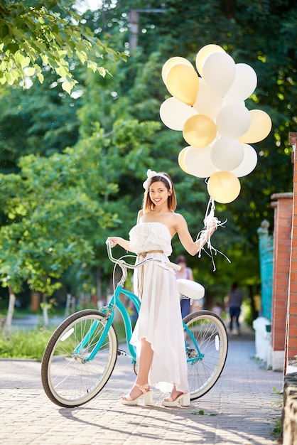 Jeune femme posant avec des ballons colorés à la main près de vélo bleu vintage sur allée du parc le jour d'été ensoleillé