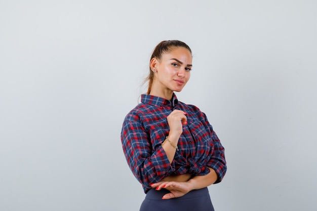 Jeune femme posant debout en chemise à carreaux, pantalon et charmante, vue de face.