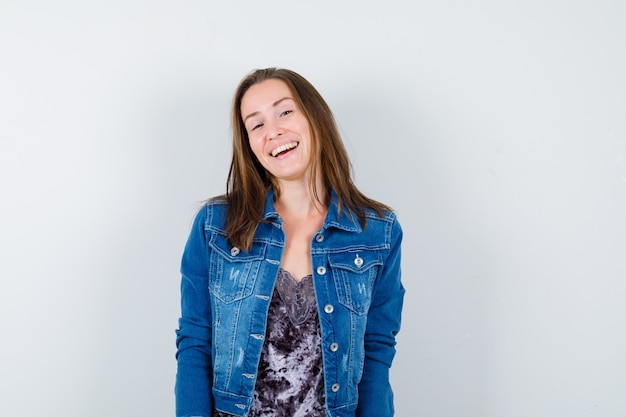 Jeune femme posant debout dans un chemisier, une veste en jean et l'air heureuse. vue de face.