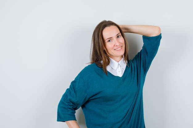 Jeune femme posant avec la main derrière la tête en chemise blanche, pull et à la joyeuse, vue de face.