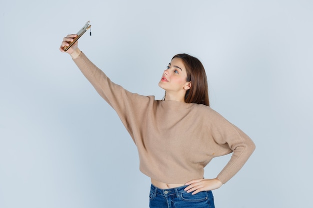 Jeune femme posant en prenant un selfie avec un téléphone portable en pull beige, jeans et semblant attrayante. vue de face.