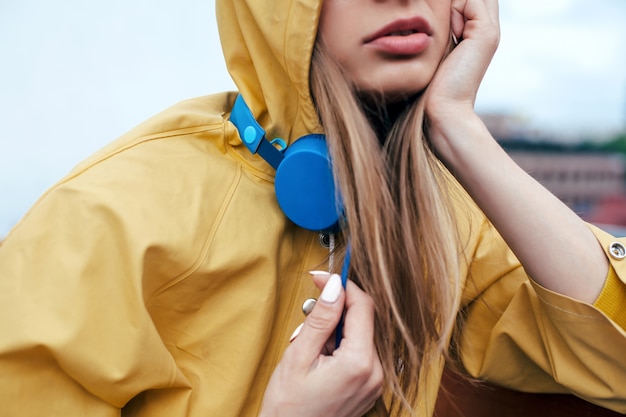 Jeune femme posent en plein air avec des écouteurs élégants bleus habillés en imperméable jaune. Closeup portrait