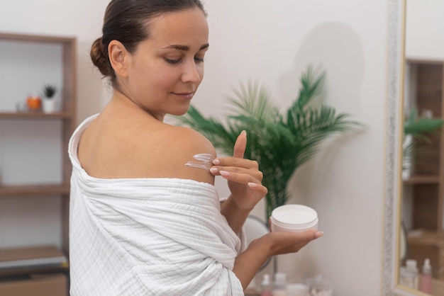 Jeune femme positive appliquant de la crème hydratante sur l'épaule nue dans les soins du corps de la salle de bain