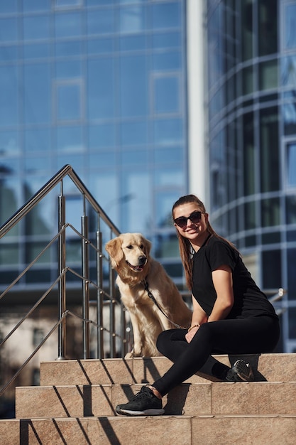 Jeune femme positive assise dans les escaliers avec son chien lorsqu'elle se promène à l'extérieur près d'un bâtiment commercial