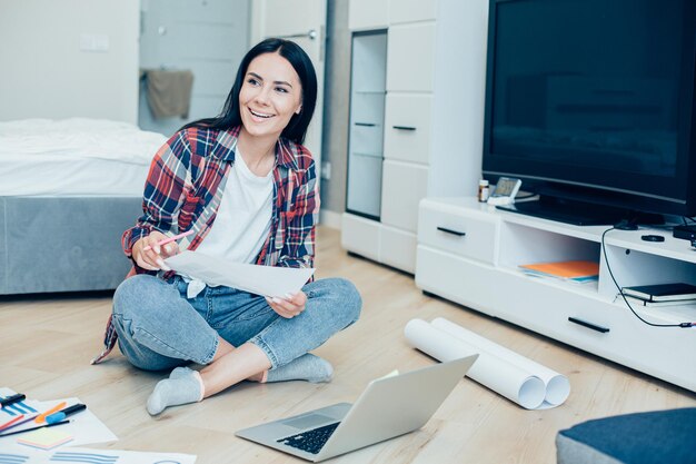 Jeune femme positive assise sur le sol avec un ordinateur portable et de nombreuses cartes graphiques et regardant au loin avec un sourire