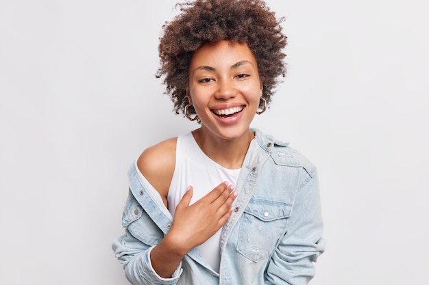 Une Jeune Femme Positive Aux Cheveux Bouclés Naturels Sourit Largement Exprime Des émotions Sincères Vêtue D'une Chemise En Jean Isolée Sur Un Mur Blanc