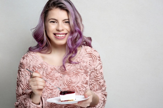 Jeune femme positive aux cheveux bouclés violets tenant un délicieux gâteau au fromage sur fond gris. Espace pour le texte