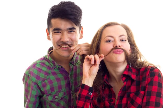 Jeune femme positive aux longs cheveux châtains et jeune bel homme aux cheveux noirs en chemises à carreaux faisant moustache de cheveux sur fond blanc en studio