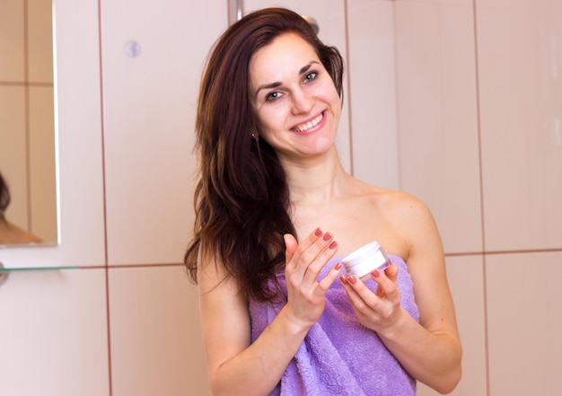 Jeune femme positive aux longs cheveux noirs dans une serviette violette tenant un pot blanc de crème dans sa salle de bain