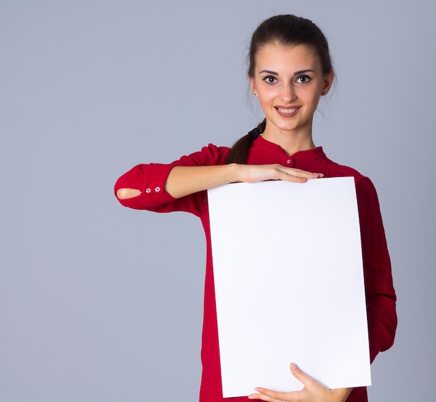 Jeune femme positive en chemisier rouge tenant une feuille de papier blanche sur fond gris en studio