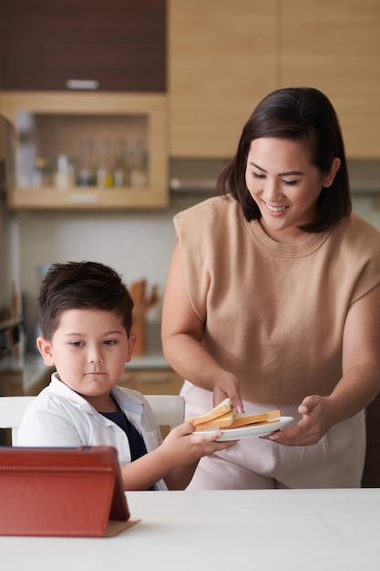 Jeune femme positive donnant une assiette de sandwichs au petit fils étudiant en ligne