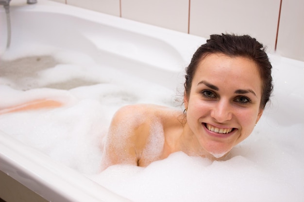 Jeune femme positive avec un paquet sur la tête se relaxant dans le bain plein de mousse dans sa salle de bain