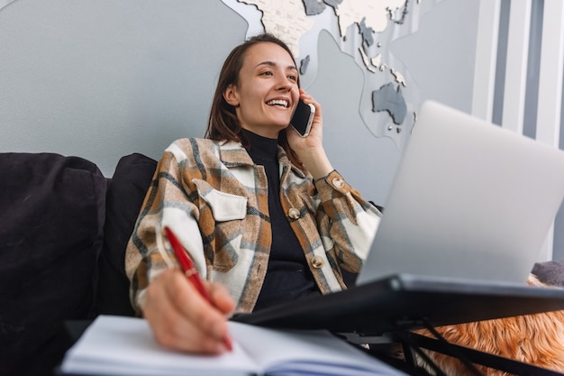 Jeune femme positive parlant au téléphone tout en travaillant sur l'ordinateur portable