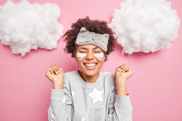 Une jeune femme positive se réjouit de bonnes nouvelles sourit largement garde les yeux fermés serre les mains dans les poings porte un pyjama et un masque de sommeil isolé sur un mur rose