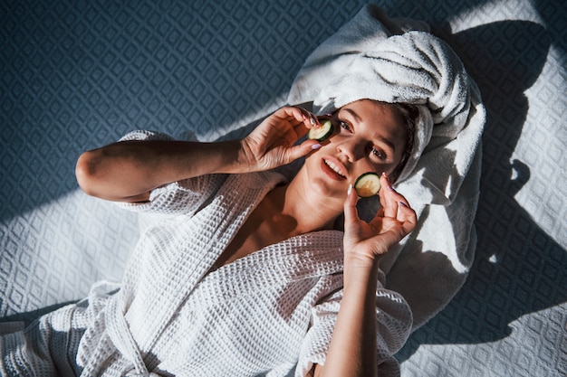 Jeune femme positive avec une serviette sur la tête allongée sur le lit avec du concombre.