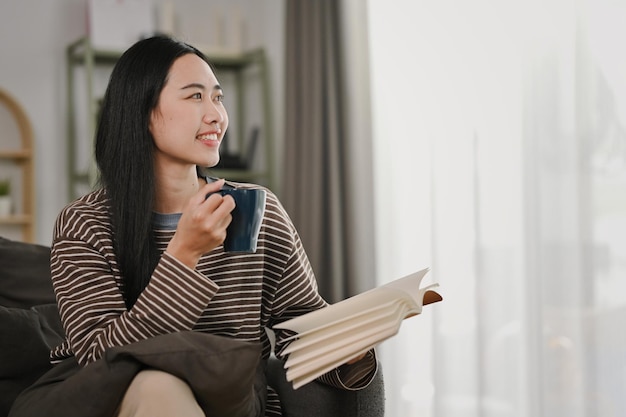 Une jeune femme positive tenant un livre et buvant du café en détournant les yeux alors qu'elle est assise dans une maison confortable