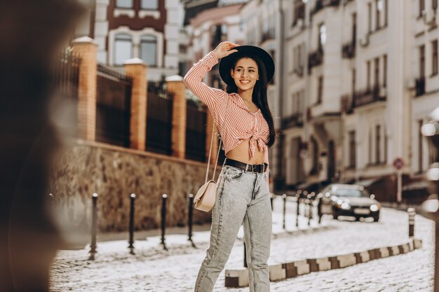 Jeune femme positive touchant son chapeau dans la rue et regardant loin avec un sourire