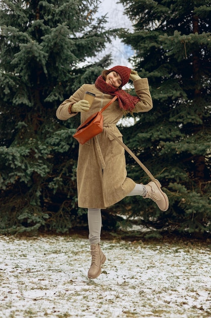 jeune femme positive en vêtements d'extérieur avec une tasse de café à emporter sautant dans un parc d'hiver enneigé