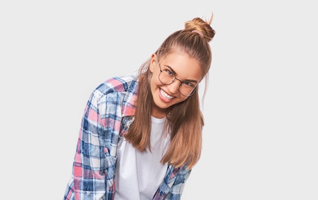 Jeune femme positive vêtue d'une tenue décontractée portant des lunettes rondes transparentes avec un large sourire agréable regardant vers la caméra et posant sur un mur de studio blanc Émotions des gens