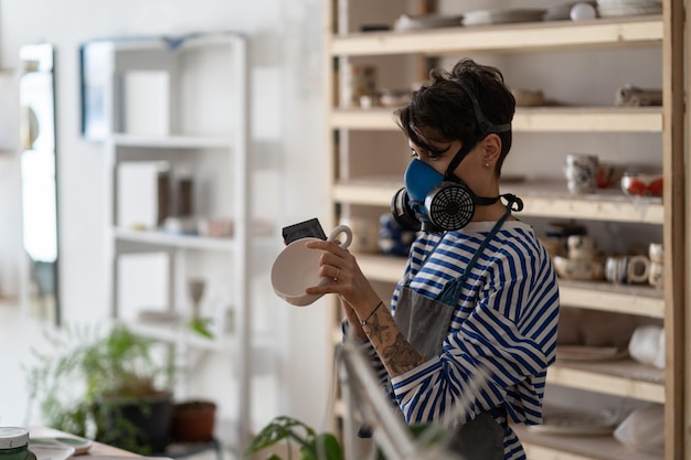 Jeune femme potier portant un respirateur ponçant la céramique avec un bloc de meulage en atelier