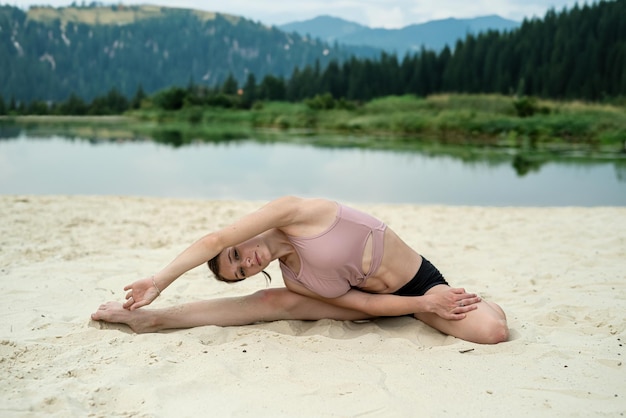 Une jeune femme pratique le yoga sur une plage de sable près d'un lac avec des montagnes en arrière-plan.