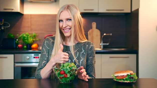 Jeune femme préférant la salade au hamburger Jolie jeune femme choisissant de manger une salade saine pour lu...