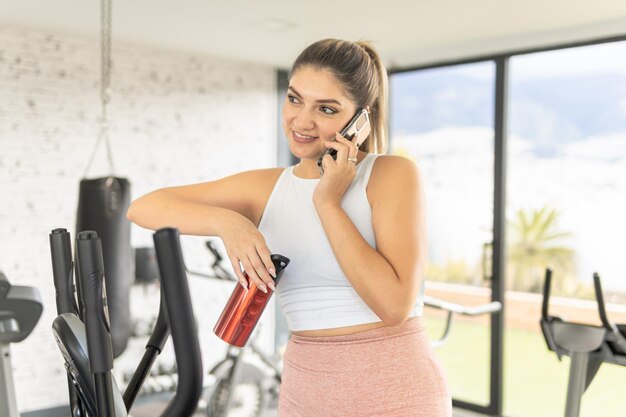 Photo une jeune femme prenant une pause en parlant au téléphone portable et en buvant de l'eau dans le gymnase