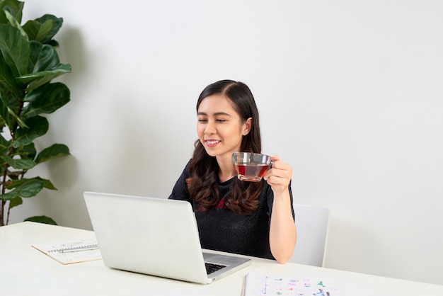 Jeune femme prenant une pause thé alors qu'elle est assise à une table travaillant sur un ordinateur portable,