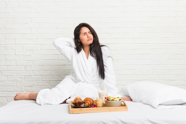 Jeune femme prenant un petit déjeuner sur le lit souffrant de douleurs au cou en raison d'un mode de vie sédentaire.