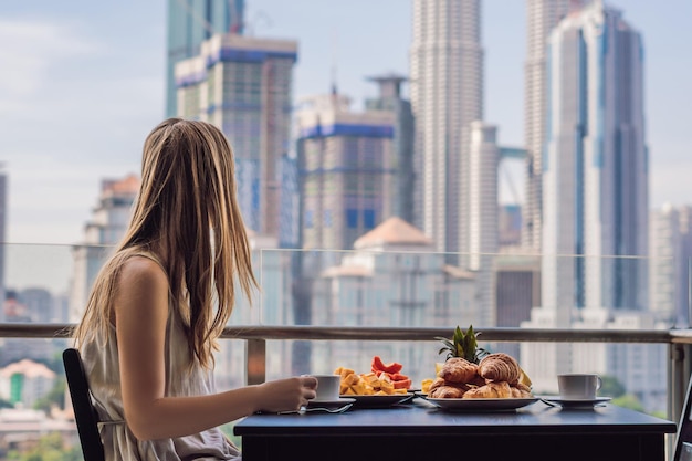 Une jeune femme prend son petit déjeuner sur le balcon. Table de petit-déjeuner avec café, fruits et pain croissant sur un balcon dans le contexte de la grande ville